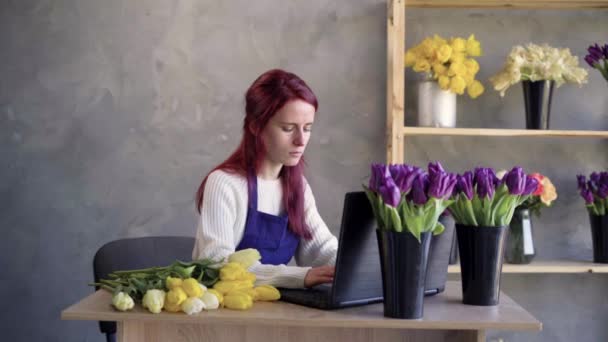 Kobieta-przedsiębiorca, kobieta-kwiaciarka pracująca na laptopie, zamawiająca rośliny. Młoda bizneswoman w fartuchu sprawdza dokumenty na komputerze w biurze z półkami z kwiatami. — Wideo stockowe