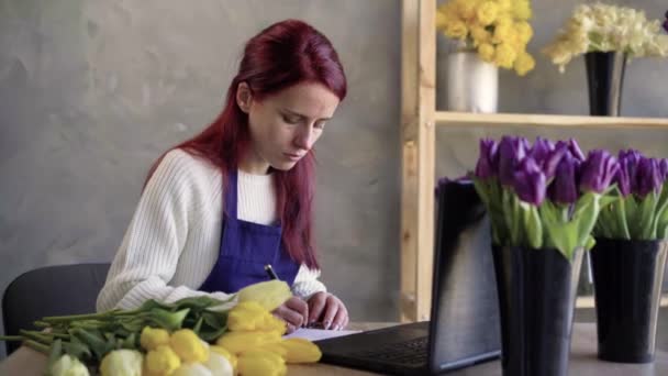 Porträt einer jungen und attraktiven kaukasischen Geschäftsfrau Floristin und Verkäuferin mit einem Laptop online bestellen, Notizen in einem Notizbuch. Arbeiten im Innenbereich. — Stockvideo