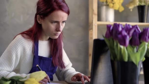 Retrato de una joven y atractiva mujer de negocios caucásica florista y vendedora utilizando un ordenador portátil para hacer un pedido en línea en línea, tomar notas en un cuaderno. Negocios de trabajo en interiores. — Vídeo de stock