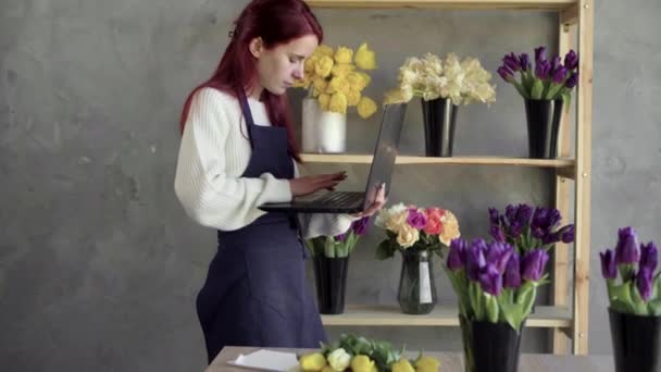 Retrato de uma florista mulher caucasiana em um avental que trabalha em sua própria loja de flores, usando um laptop e escrever uma ordem para um buquê. fica perto da vitrine com flores e um sorriso confidencial — Vídeo de Stock