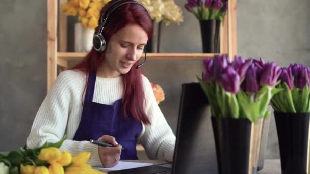 Florista en auriculares con un micrófono y un delantal se sienta en una mesa, utiliza un ordenador portátil para el trabajo, videollamada en línea. Trabajo desde casa. El operador organiza logística y entrega, toma pedidos. — Vídeo de stock