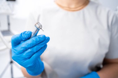 Mavi eldivenli bir dişçinin elinde bir alet, bir matkap var. Parlak bir kliniğin arka planında dişçi aleti var. Yakın plan..