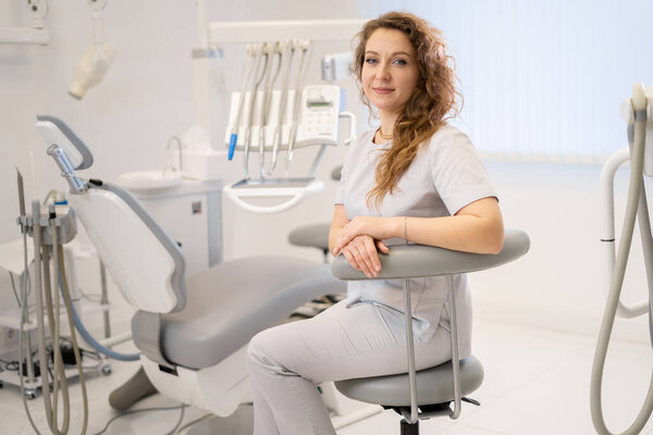 Beautiful young dentist woman sitting on a chair in her clinic smiling at the camera, dentistry and medicine. copy space