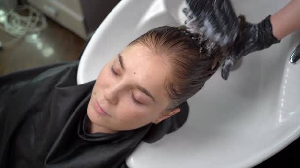 Jeune femme, couchée dans un salon, se lave les cheveux dans un salon de coiffure par un styliste. Beau client relaxant tout en coiffeur massant et lavant les cheveux avec de l'eau et du shampooing — Video