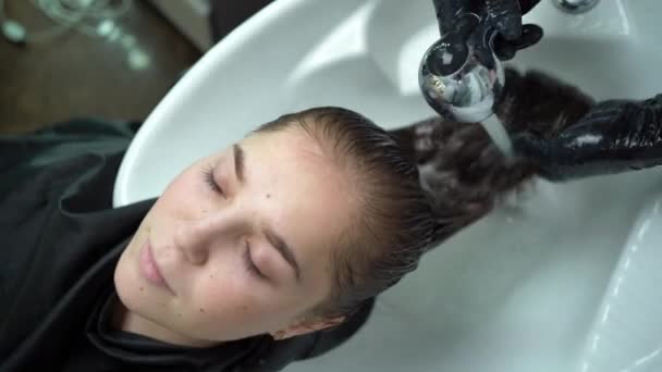 Jovem, encontra-se em um salão de beleza, lava o cabelo em um salão de cabeleireiro por um estilista. Cliente bonito relaxante enquanto cabeleireiro massagear e lavar o cabelo com água e xampu — Vídeo de Stock