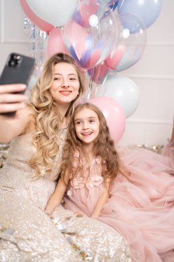 Hassas pembe elbiseler giymiş bir kadın ve bir çocuk helyum balonlarının arka planında selfie çekiyorlar. Boşluğu kopyala.