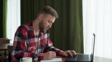 Odaklanmış beyaz bir adam masaya oturur, bir web sitesi izler, dizüstü bilgisayar kullanır, yeni bilgiler kazanır, notlar yazar. Video konferans iletişimi, uzaktan müzakere, çevrimiçi öğrenme, e-öğrenme,