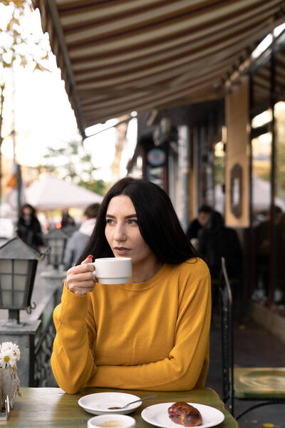 кавказская стильная женщина, модный свитер, сидя в кафе, держа чашку капучино, наслаждаясь брюнеткой 30-х годов, с днем рождения, улица города, бохо наряд, европейский отпуск, романтический ужин, солнечный