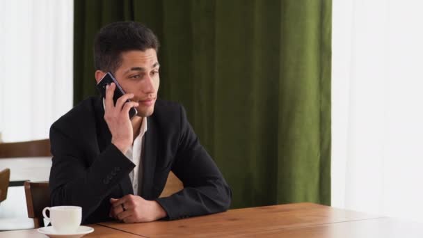 Retrato de um alegre árabe millennial cara conversando em um smartphone, sentado à mesa em um apartamento, um homem positivo de bom humor falando ao telefone, falando em um telefone celular em uma jaqueta — Vídeo de Stock