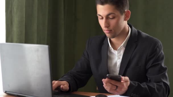 Biznesmen w kurtce pracuje w biurze, siedzi przy stole, wprowadza dane ze smartfona do laptopa. Arab pracuje zdalnie.. — Wideo stockowe