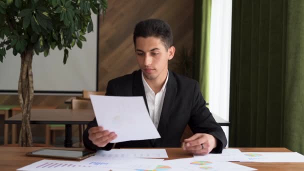 Συγκεντρωμένος νεαρός Άραβας επιχειρηματίας που εξετάζει την έκθεση έρευνας μάρκετινγκ, αναλύει στατιστικά δεδομένα σε διαγράμματα, αναπτύσσει στρατηγική ανάπτυξης, εργάζεται μόνος του στο σύγχρονο γραφείο — Αρχείο Βίντεο