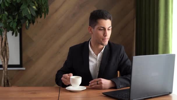 Jovem homem árabe usando computador portátil, sentado à mesa no escritório em casa, bebendo café lendo notícias da manhã, desfrutando de trabalho remoto — Vídeo de Stock