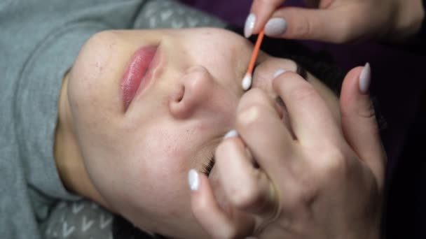 Eine junge Frau unterzieht sich einer Wimpernverlängerung und entfernt Mascara mit Wattestäbchen und Stick in einem Schönheitssalon. Augenlidpräparation. — Stockvideo