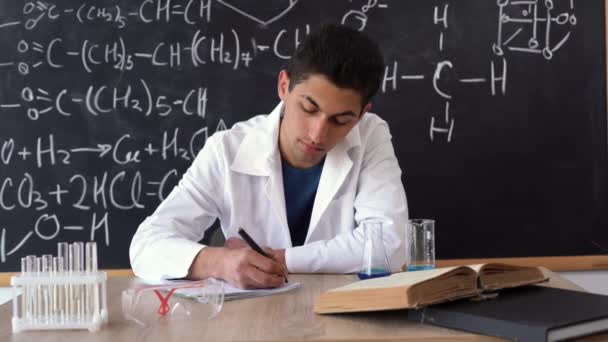 Ein junger Wissenschaftler Chemiker an der Universität bereitet sich auf eine Prüfung vor, studiert Chemie als Naturwissenschaft in der Schule, Arabisch Student in einer Chemiestunde, Tafel Hintergrund mit chemischen Formeln — Stockvideo