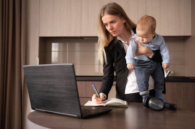 Ofiste küçük bir bebekle çalışan genç beyaz bir kadın. Anne oğlunu kollarına aldı, dizüstü bilgisayarın yanında durdu ve not defterine not aldı. Boşluğu kopyala.