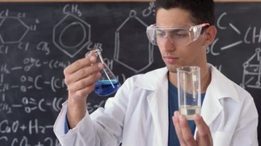Arap görünümlü bir erkek bilim adamı laboratuvarda kimyasal formüllü bir panonun arka planına karşı iki test tüpü tutuyor, araştırma ve analiz yapıyor..