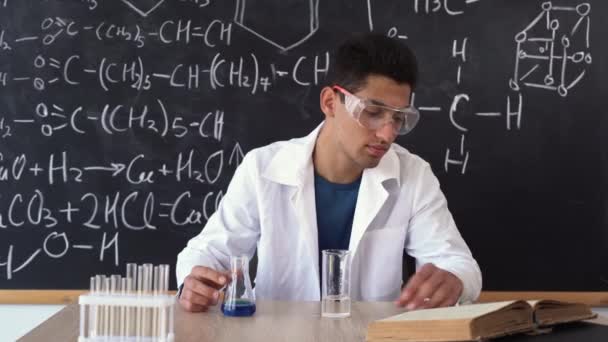 Un homme arabe est assis dans le public à une table sur le fond d'un tableau noir avec des formules chimiques, lit un livre et regarde une éprouvette avec un liquide — Video