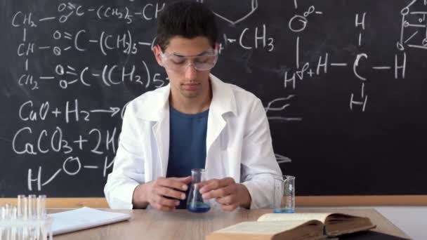 Un insegnante di chimica araba maschile in bicchieri siede a un tavolo mescola un liquido colorato in un bicchiere da laboratorio. Concentrati sulle mani. — Video Stock