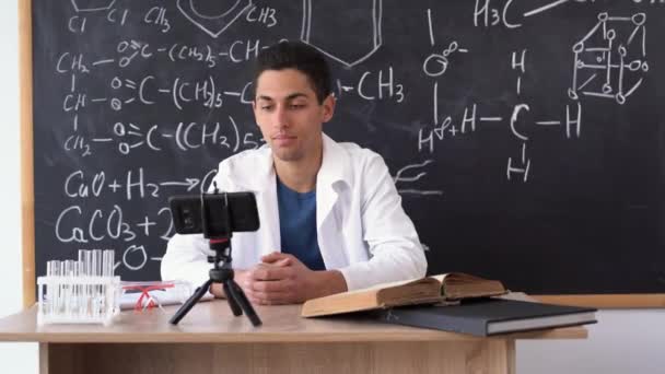 Νεαρός καθηγητής σύγχρονης Αραβικής Χημείας με λευκό παλτό κατά τη διάρκεια ενός online μαθήματος ενάντια σε έναν πίνακα με χημικούς τύπους. — Αρχείο Βίντεο