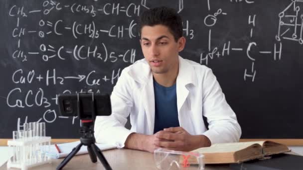 Молодой учитель современной арабской химии в белом халате во время онлайн-урока против доски с химическими формулами. — стоковое видео