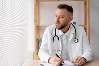 Kafkasyalı bir doktor, beyaz önlüklü, parlak bir ofiste, hastane günlüğünde steteskop bulunan bir masada oturuyor. Tıp, sağlık hizmeti kavramı.