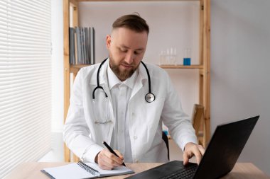 Ciddi bir erkek doktor masada otururken dizüstü bilgisayar kullanıyor ve bir tıp dergisine notlar yazıyor. Beyaz önlük ve steteskopla işyerindeki bilgisayarda çalışıyor..