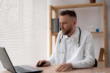 Beyaz önlüklü ve steteskoplu bir erkek doktor hastanedeki bir masada oturuyor ve dizüstü bilgisayarla yazı yazıyor..