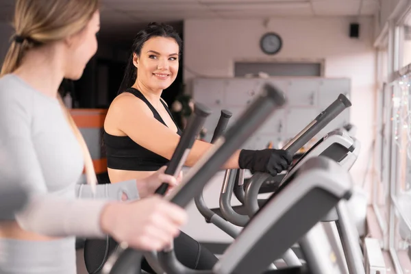 Twee aantrekkelijke atletische jonge vrouwen trainen samen in de sportschool. Uitvoeren van oefening op cardiomachine. — Stockfoto