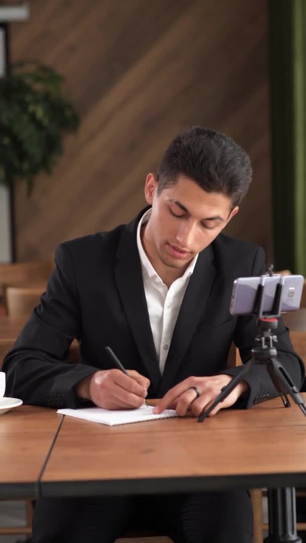 Studente universitario arabo felice o insegnante con smartphone sul bancone con un sorriso carino, annotando una lezione da un gadget elettronico in un quaderno, seduto a un tavolo in un caffè — Video Stock