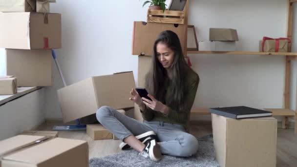 Mulher 25s sentado no chão com um monte de caixas, usando um telefone celular, encomendar através de um site para mover serviços no dia em movimento, comprar móveis on-line — Vídeo de Stock