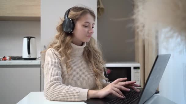 Genç ve çekici bir kadın mutfakta uzaktan bilgisayarla çalışıyor, kulaklık takıyor ve gülümsüyor.. — Stok video