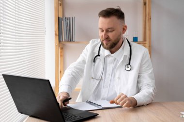 Ciddi bir erkek doktor masada otururken dizüstü bilgisayar kullanıyor ve bir tıp dergisine notlar yazıyor. Beyaz önlük ve steteskopla işyerindeki bilgisayarda çalışıyor..