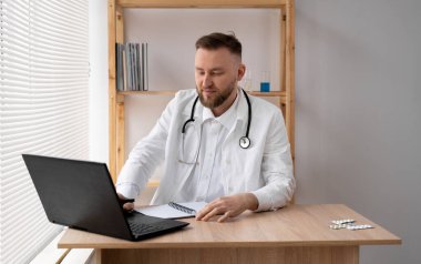 Steteskoplu beyaz önlüklü genç bir doktorun portresi, bir tıp ofisinde oturuyor ve dizüstü bilgisayarda çalışıyor. Boşluğu kopyala.