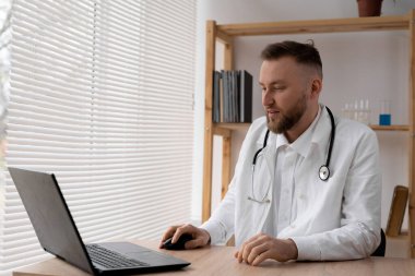 Beyaz önlüklü ve steteskoplu bir erkek doktor hastanedeki bir masada dizüstü bilgisayarla bilgi arıyor. Boşluğu kopyala.