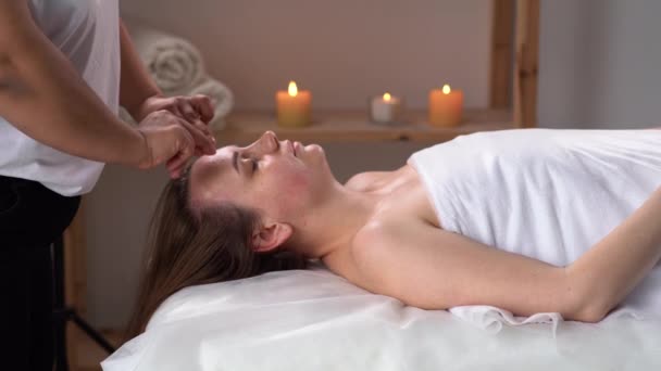 Acupressure facial massage for a young woman in a spa massage salon, side view. spa attendant making relaxing massage for woman wrapped in white towel pressing on her forehead — Stock Video