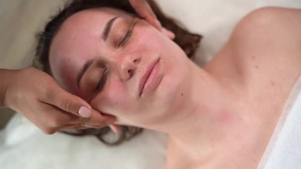 Rahatlamış genç bir kadın lüks spa merkezindeki masör tarafından yüz masajı ve kafa masajı için spa yatağında yatıyor. Sağlık, stres rahatlama kavramı — Stok video