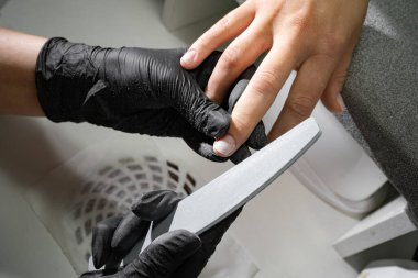 Eldiven giymiş deneyimli bir manikürcünün elleri kaplıca salonunda manikür yaparken tırnak törpüsü takan genç bir kadının tırnaklarını törpülüyor. yakın plan.