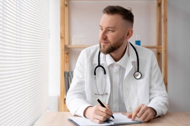 Hastanedeki bir masada oturan beyaz üniformalı ciddi bir pratisyen hekimin portresi, steteskoplu bir doktor ya da terapist iş, tıp ve sağlık hizmetleri konseptine pencereden bakar.