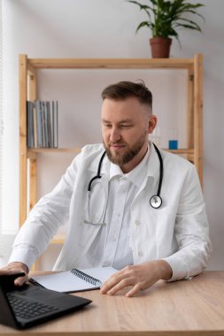 Steteskoplu beyaz önlüklü genç bir doktorun portresi, bir tıp ofisinde oturuyor ve dizüstü bilgisayarda çalışıyor..