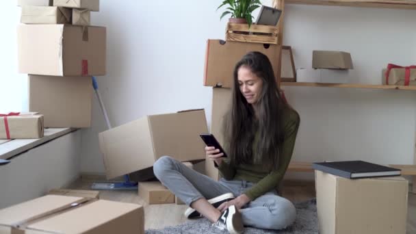 Nieuw huis, aantrekkelijke jonge latijnse vrouw in beweging, zittend tussen kartonnen dozen, met behulp van smartphone en glimlachen, verhuizen en relocatie — Stockvideo