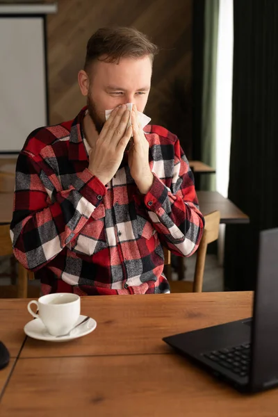 Ill caucasian man looking at laptop screen and sneezing nose in paper tissue, sitting on the office, empty space. Sinusitis illness, cold and influenza symptoms, allergy, air conditioner disease