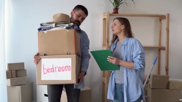 Zamilovaný pár v novém prázdném bytě stojí. Muž drží krabici s věcmi, žena zaznamenává přítomnost v seznamu věcí. — Stock video