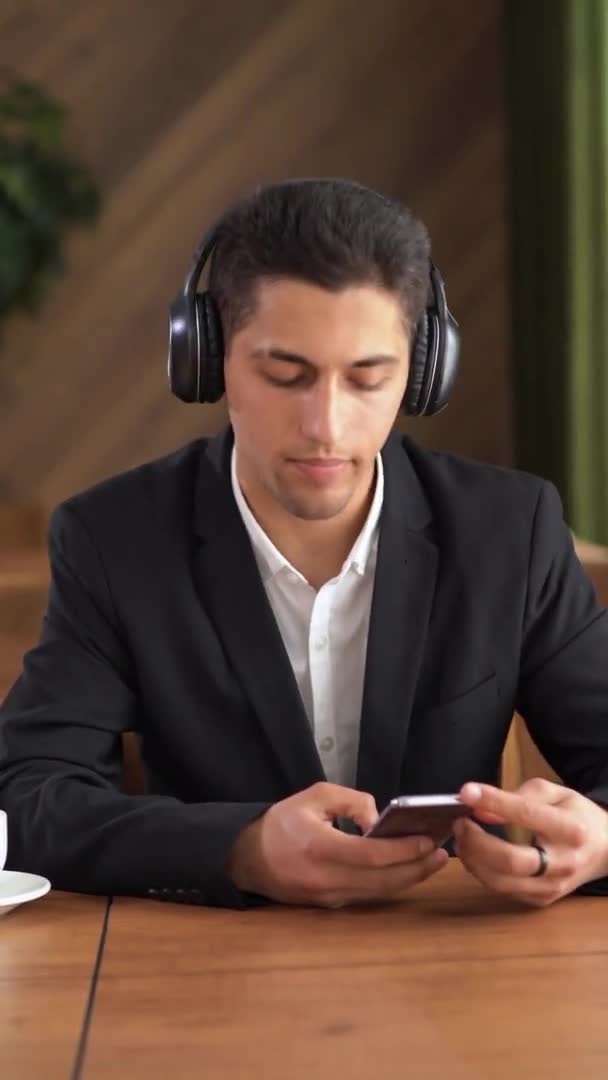 Arabe homme aime écouter de la musique par écouteurs. Il s'assoit en dansant et agitant les bras. Il est assis à la table. Homme d'affaires pendant une pause jouissant d'une mélodie à l'aide d'un smartphone — Video