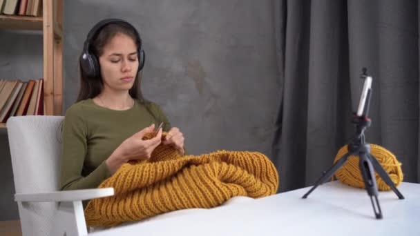 Mujer moderna en auriculares está tejiendo en casa gracias a contenido de video tutorial en línea en el teléfono inteligente y conexión inalámbrica. — Vídeos de Stock
