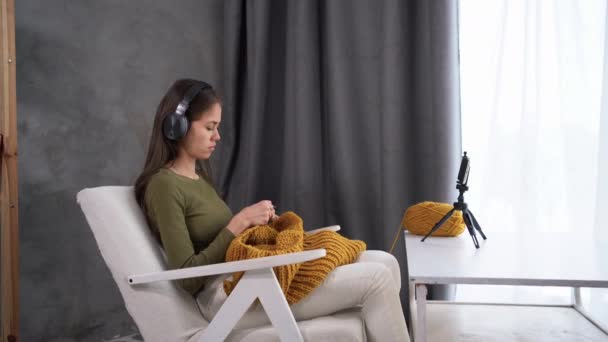 Μια σύγχρονη γυναίκα με ακουστικά ασχολείται με το πλέξιμο στο σπίτι χάρη στο να κάθεται σε μια καρέκλα και να βλέπει μια ταινία ή μια σειρά online χρησιμοποιώντας ένα smartphone. — Αρχείο Βίντεο