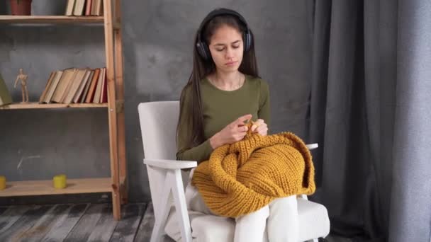 Girl in headphones knits an orange sweater at home while sitting in chair. Zero Waste Handmade, Upcycling, New Small Business Job Opportunity Concept. Hobby knitting and needlework for mental health — Vídeos de Stock
