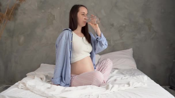 Lifestyle portrait of a young pregnant woman sitting on the bed and drinking water from a glass after waking up. Caucasian model relaxing in the morning at home. Copyspace. — Vídeo de Stock