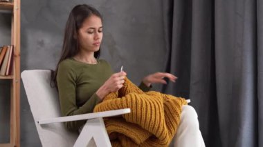 Hobi ve dikiş konsepti, genç Latin bir kadının evde koltukta oturup kalın kazak örmesi, yün ürünleri.