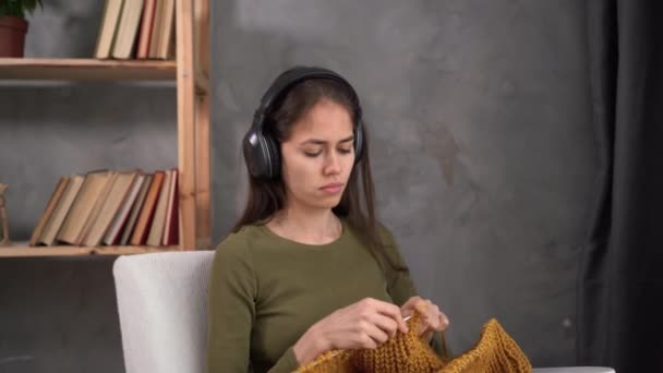 Online video knitting lesson, young latin woman knits a sweater sitting in a chair with headphones listening to an educational video, leisure needlework, hobby — 비디오