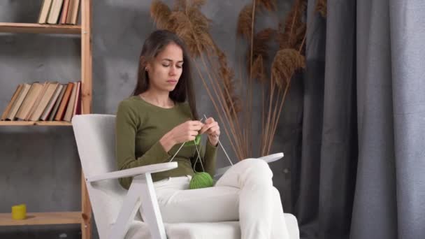 Mujer latina enfocada sosteniendo agujas sentadas en un sillón y disfrutando tejiendo. Muchacha de punto bufanda hecha a mano en casa, hobby hecho a mano — Vídeos de Stock